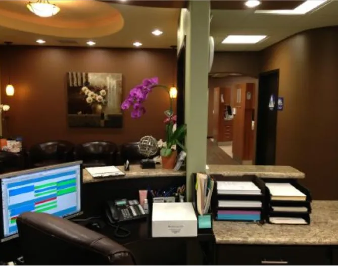 The front desk area: a reception counter in dark wood and granite, leather waiting chairs, an orchid, and a corridor leading to the treatment rooms.