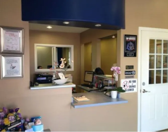 A reception window with a deep navy soffit above it, framed certificates on the wall, flowers on the counter, and a white door to the treatment area.